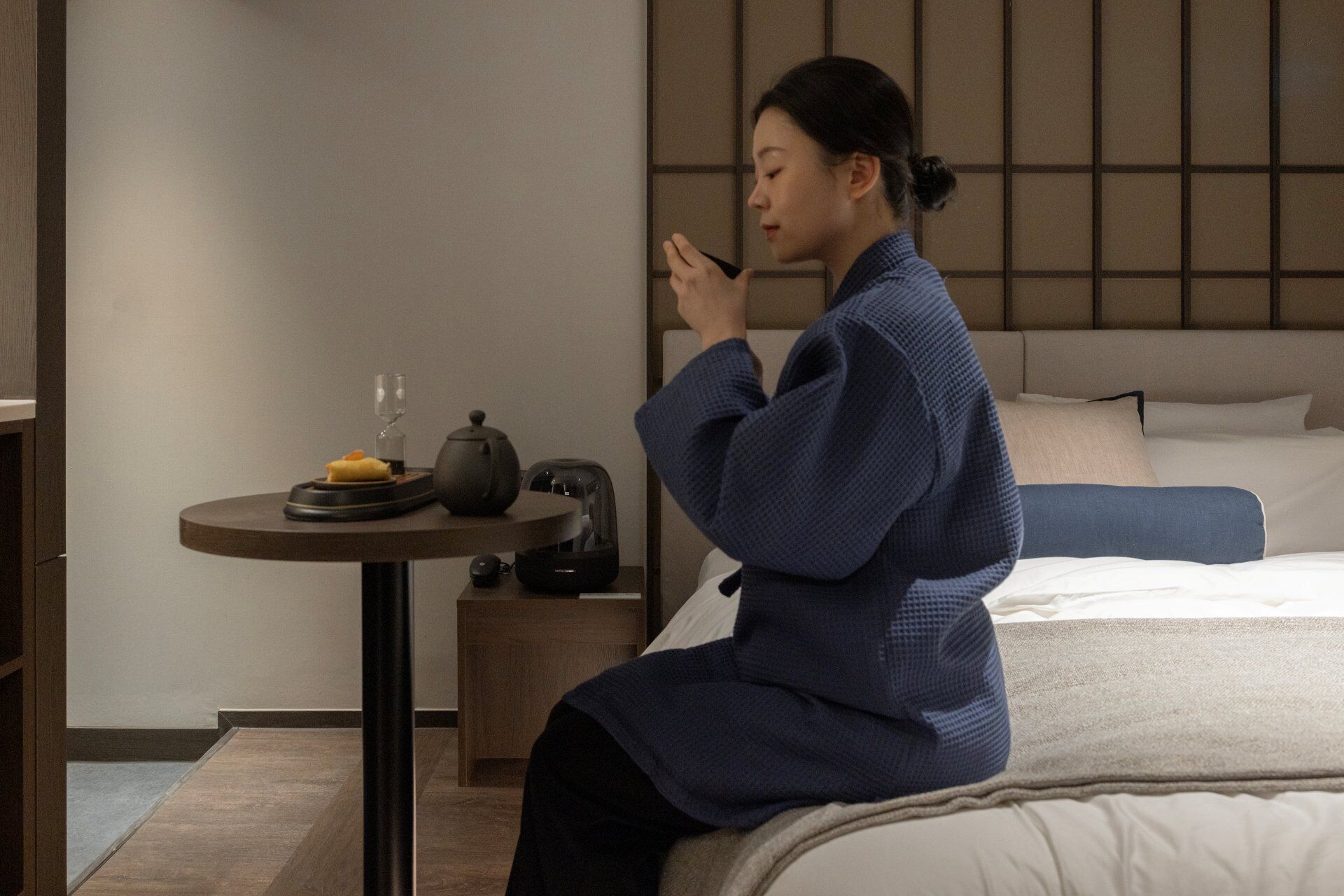 A person sitting on a bed in a room with a small table, a nightstand, and a headboard.