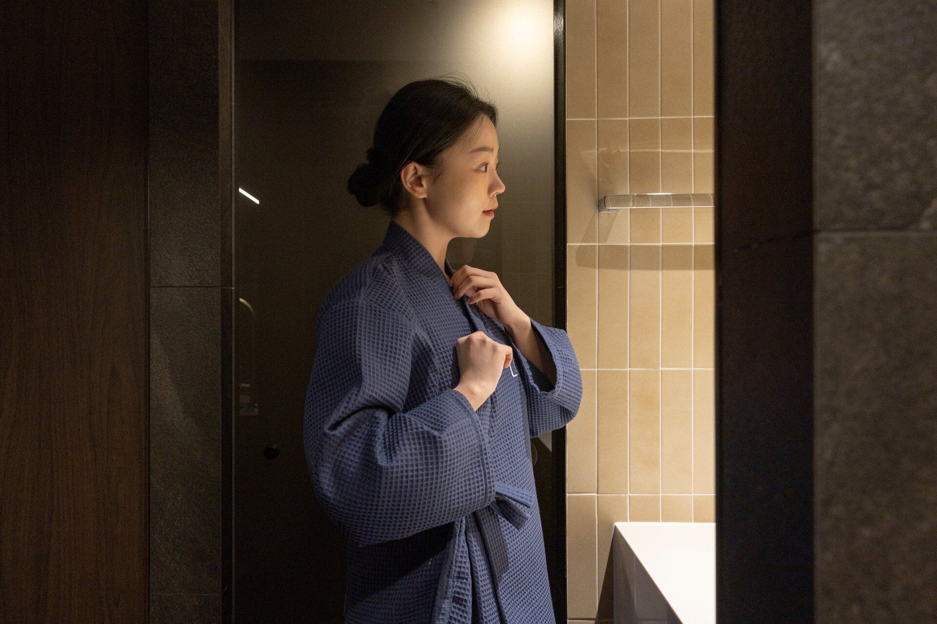 Una persona in accappatoio in un bagno con porta in vetro e pareti piastrellate.