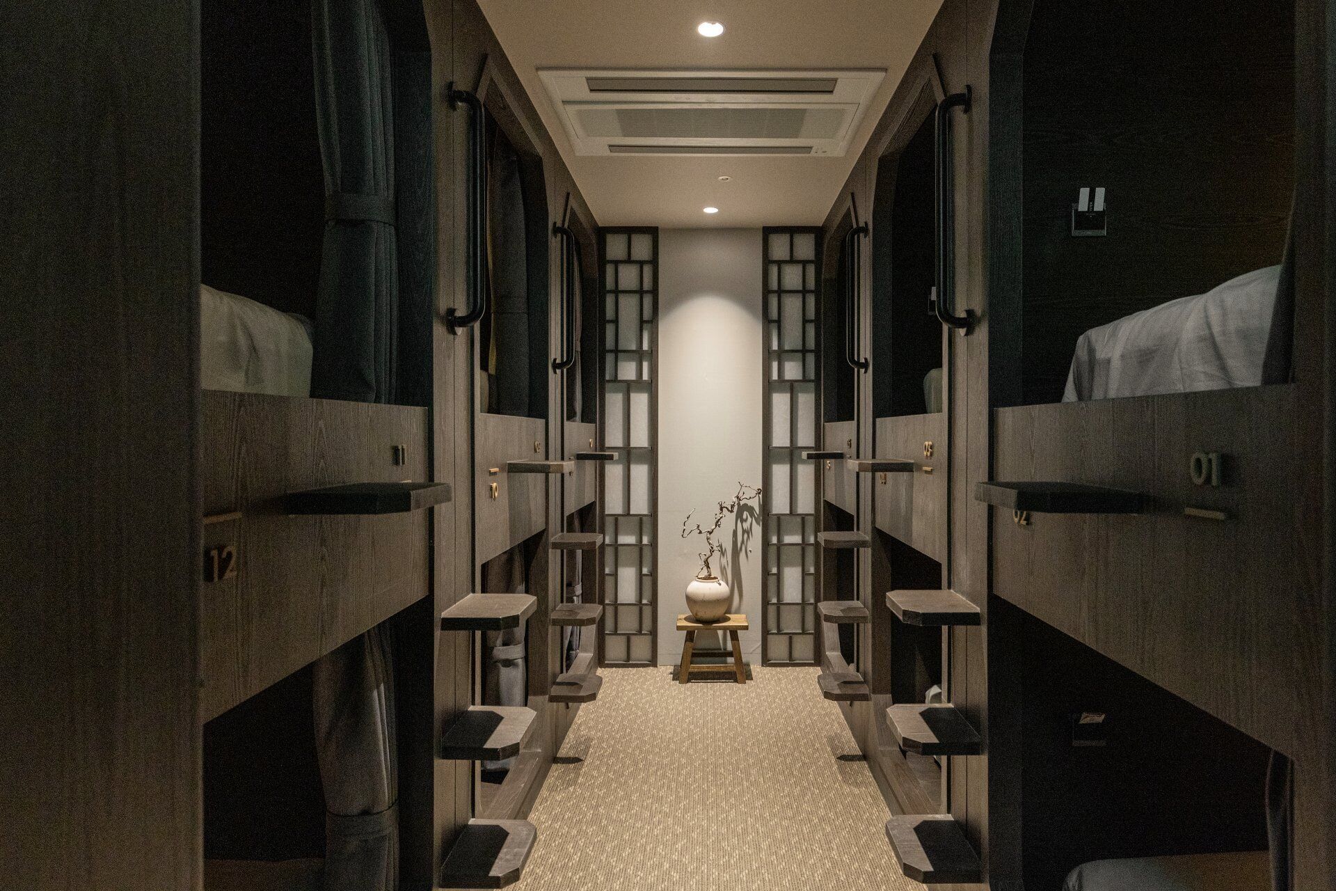 A narrow hallway with bunk beds on either side, a central light fixture, and a small table with a vase.