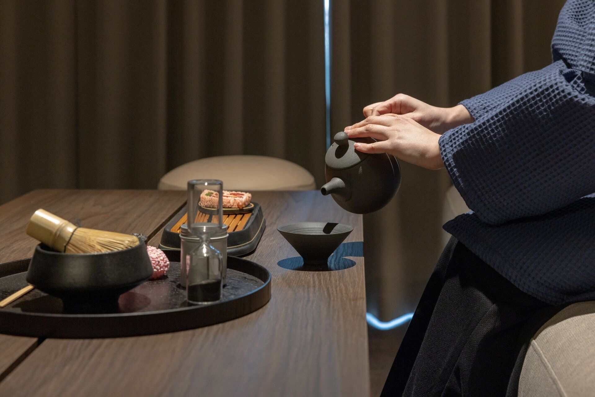 A person pouring tea from a teapot into a cup, with a tray containing a whisk, a glass, and a bowl on a wooden table.