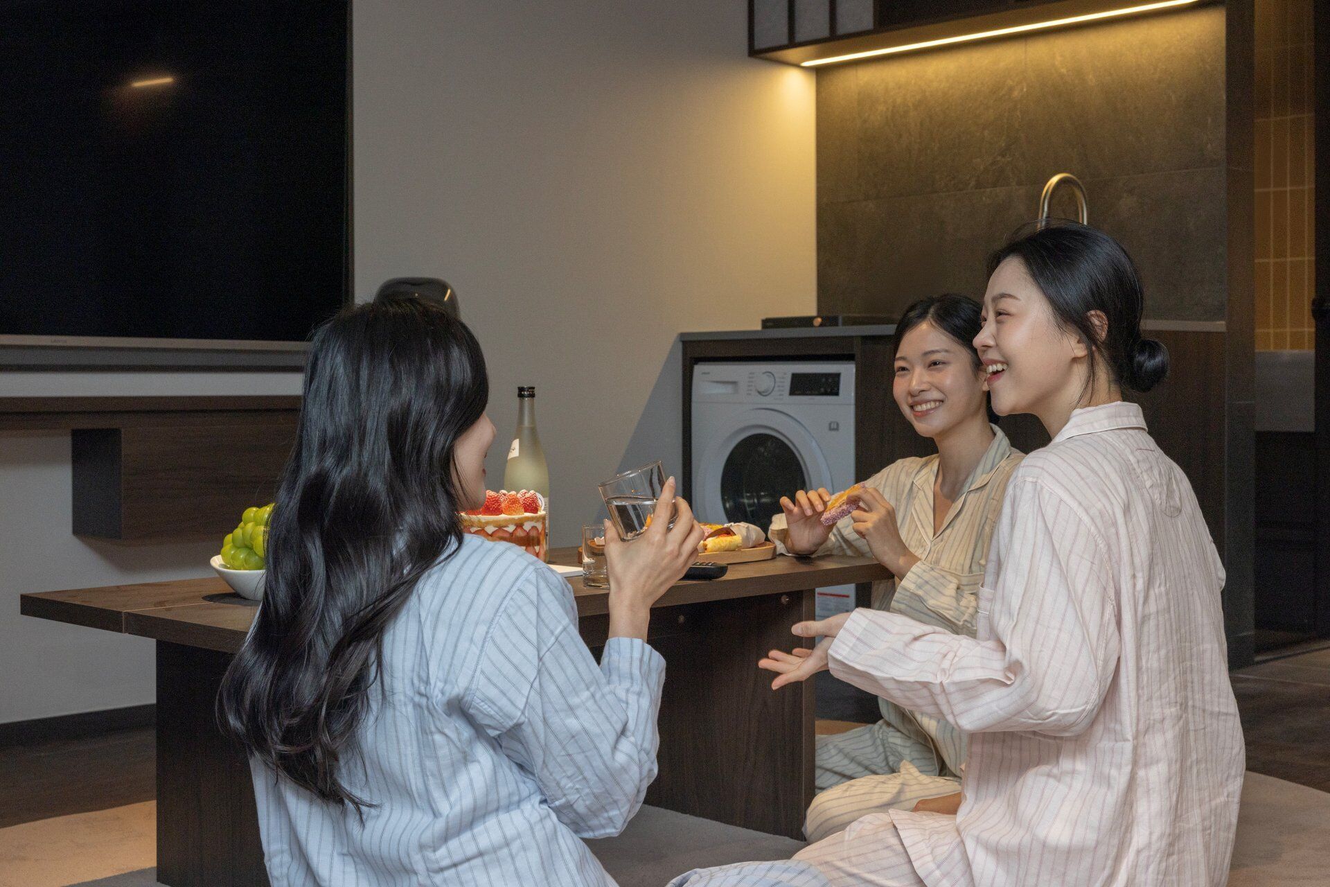 Une cuisine moderne équipée d’un lave-linge, d’une table à manger avec une bouteille et des verres, et d’un saladier rempli de fruits.