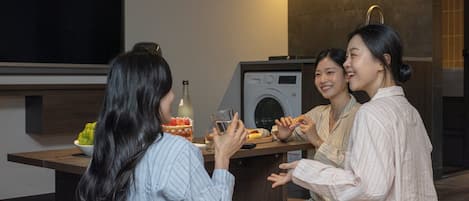 A modern kitchen with a washing machine, a dining table with a bottle and glasses, and a bowl of fruit.