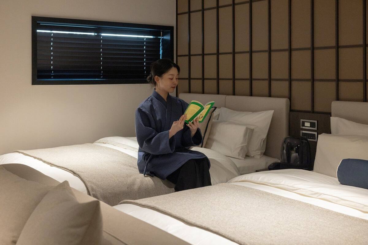 Ein Hotelzimmer mit zwei Betten, einem Fenster mit Jalousien, einer Wand mit Rillenmuster und einer Person, die ein Buch liest.
