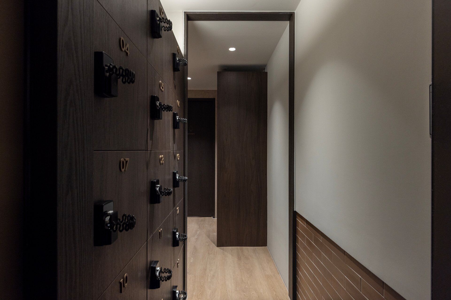A hallway with a row of dark wooden lockers on the left, a wooden floor, and a plain wall on the right.