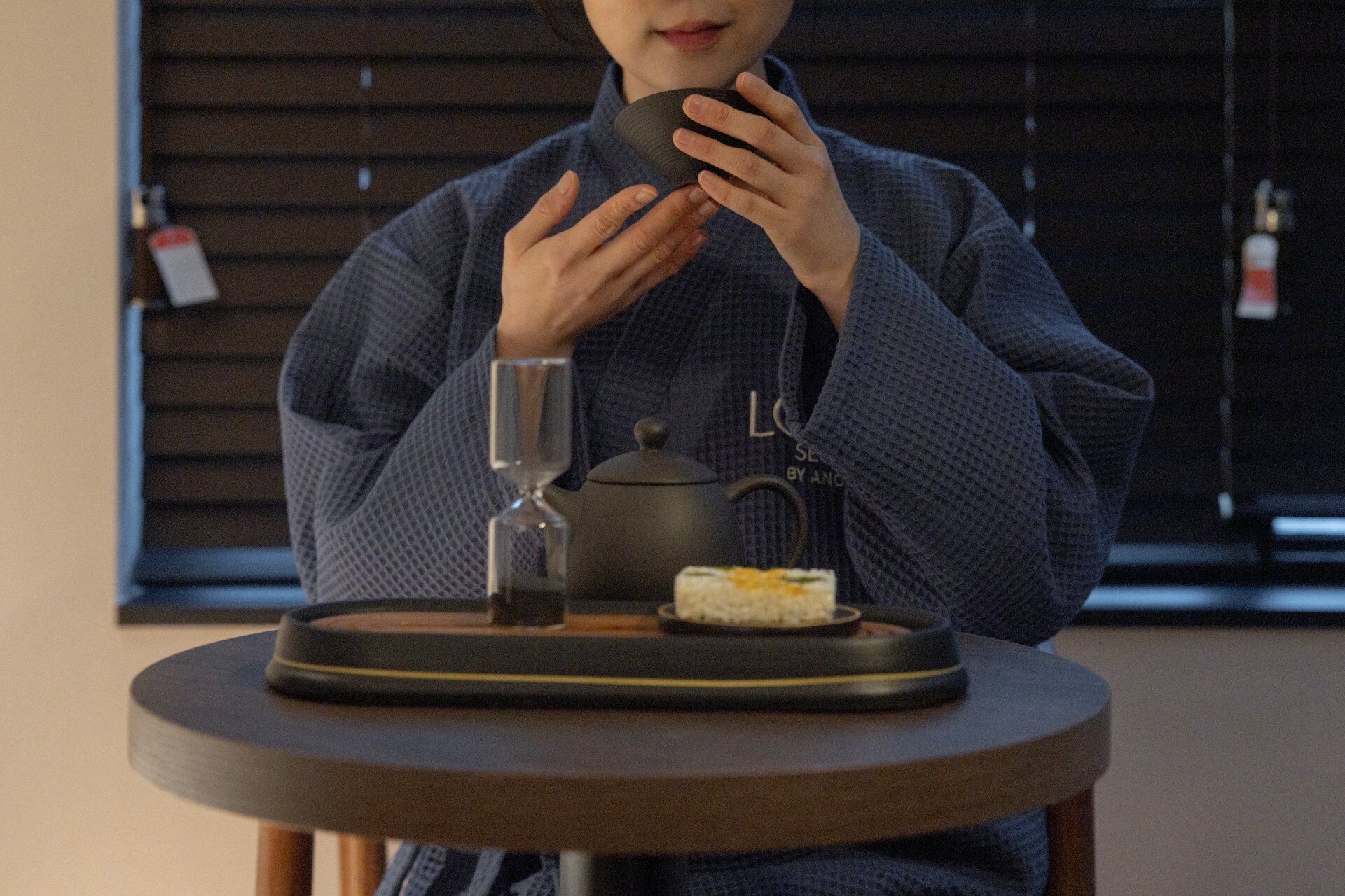 Una persona sosteniendo una taza, sentada en una mesa con una tetera, un vaso y un plato de comida.