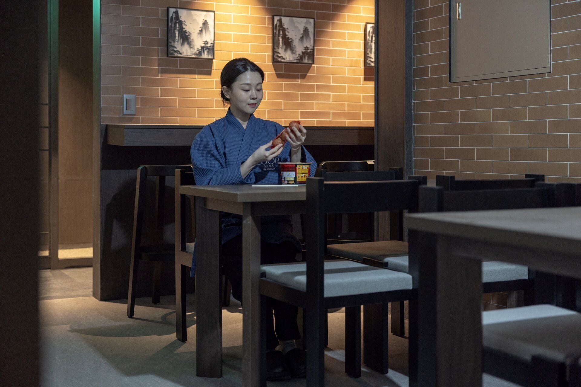 Une personne est assise à une table, devant une boisson et une boîte, dans une pièce aux murs en briques et ornée de tableaux encadrés.