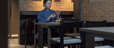 Une personne est assise à une table, devant une boisson et une boîte, dans une pièce aux murs en briques et ornée de tableaux encadrés.