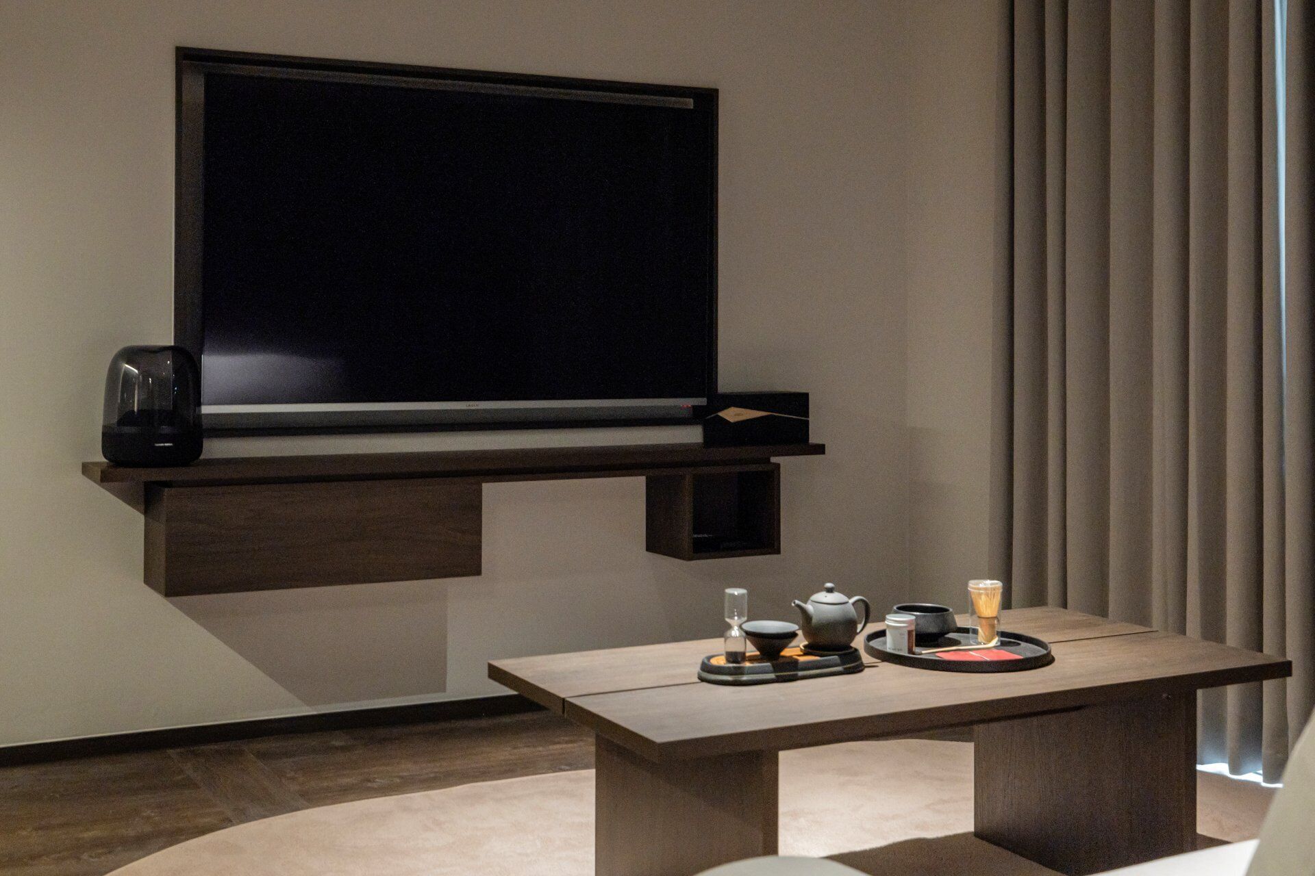 A modern room with a flat-screen TV mounted on a dark wooden shelf, a small table with a teapot, cups, and a tray, and a large window with vertical blinds.