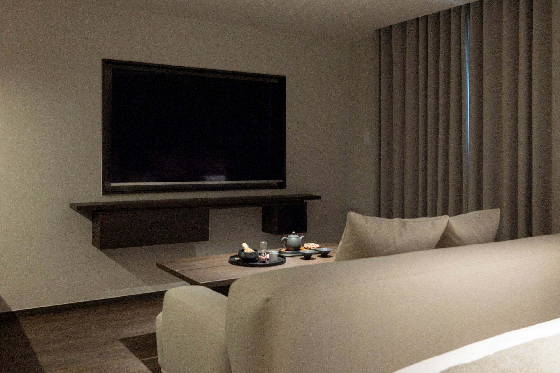 A hotel room with a flat-screen TV mounted on the wall, a small table with a teapot and cups, and a sofa with pillows.