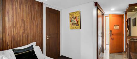 Quarto de hotel com cabeceira de madeira, cama com roupa de cama branca, porta de madeira e corredor que leva a outro quarto.