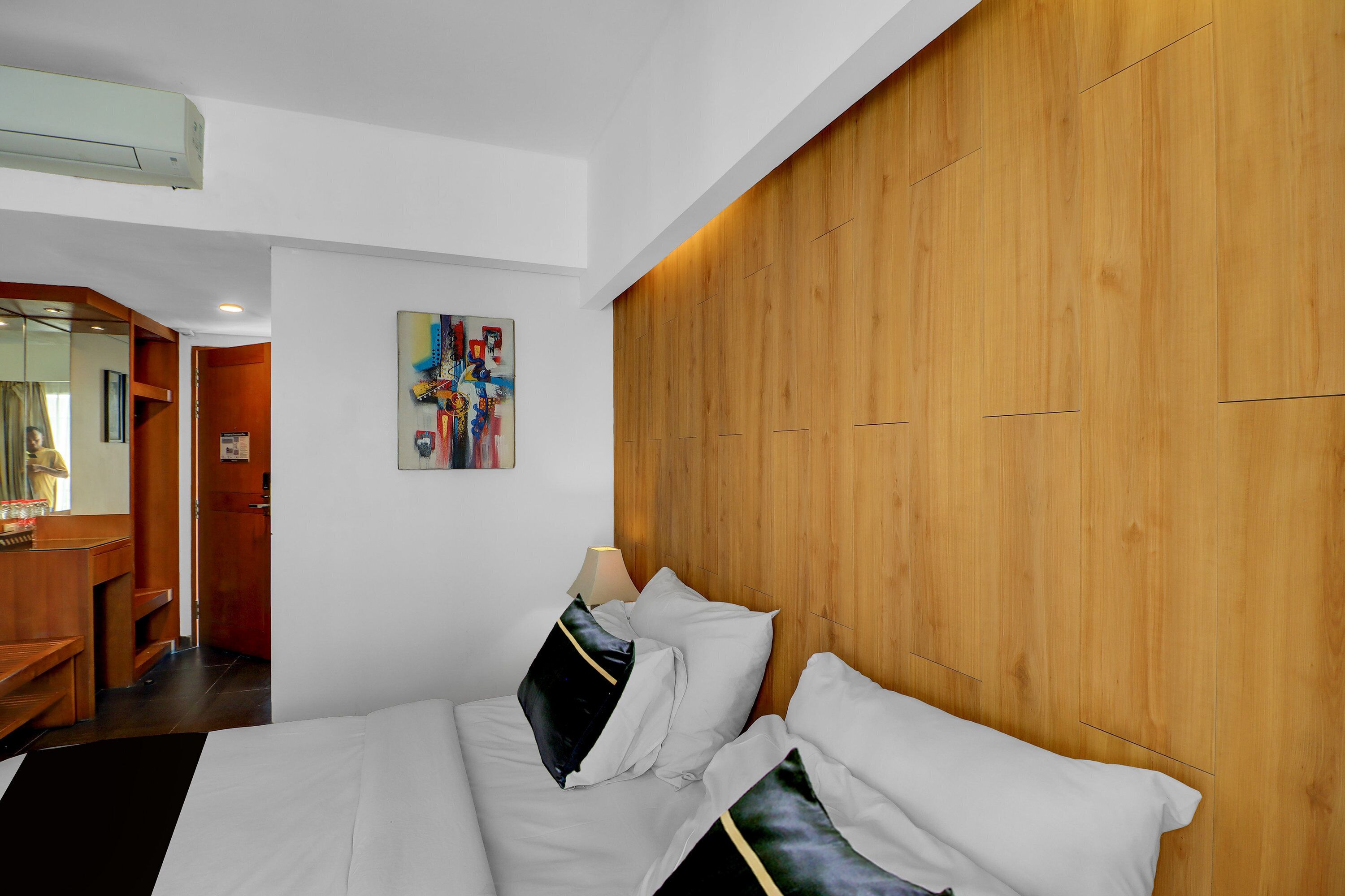 A hotel room with a wooden headboard, a bed with white bedding, a nightstand, a lamp, and a painting on the wall.