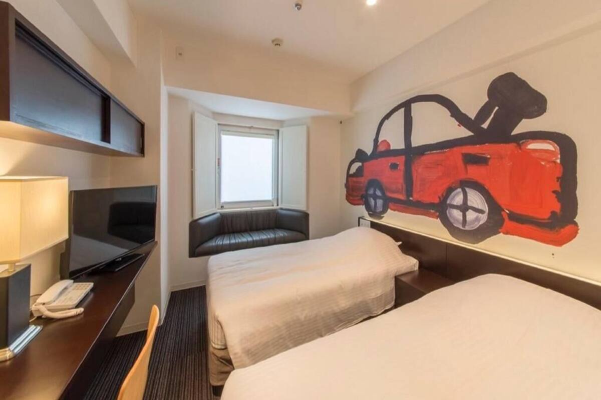 Ein Hotelzimmer mit zwei Betten, einem Schreibtisch mit Lampe, einem Fernseher und einem großen Gemälde eines Autos.