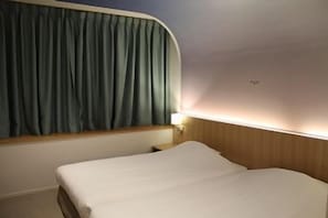 Uma cama de solteiro com cabeceira de madeira, uma janela com cortinas e um ponto de luz fixado na parede.