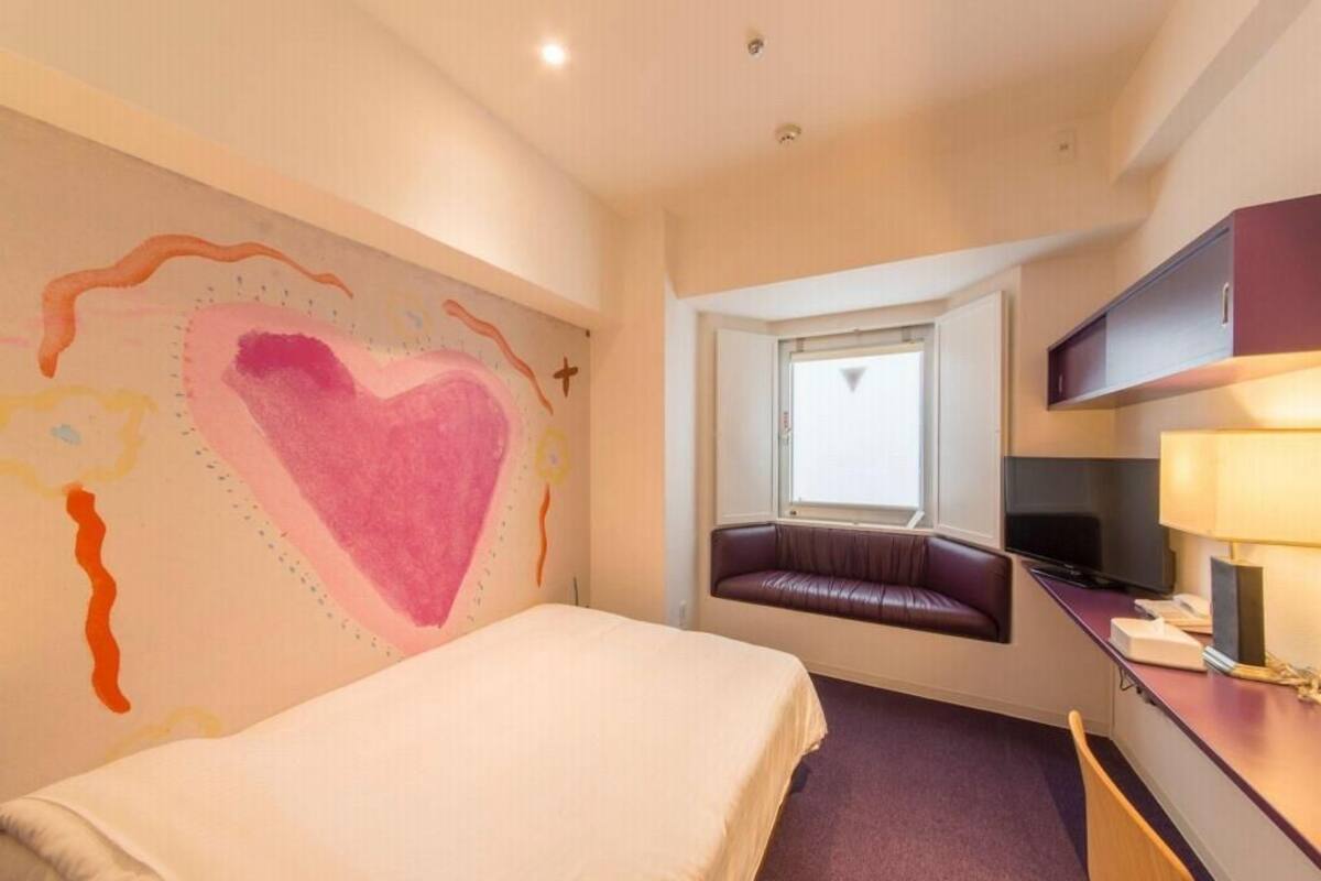 Ein Hotelzimmer mit Bett, Schreibtisch mit Lampe, Fernseher, Fenster mit Blick und einer Wand mit herzförmigem Dekor.