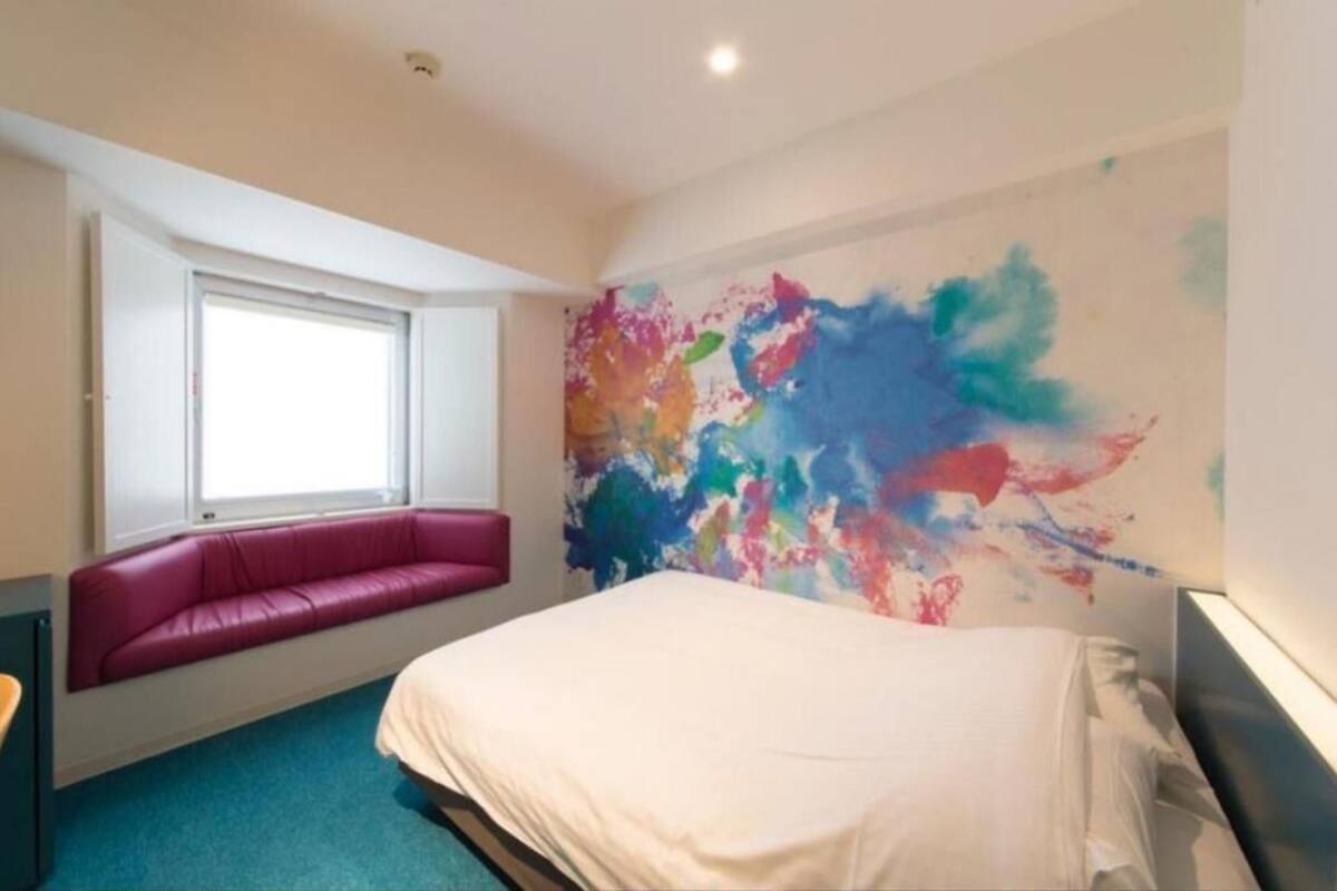 Ein Hotelzimmer mit einem Bett, einem Fenster, einer farbenfrohen abstrakten Wand und einer pinken Bank.