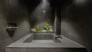 Un baño moderno con una bañera rectangular con mampara de vidrio, paredes de piedra oscura y un estante con artículos de aseo.