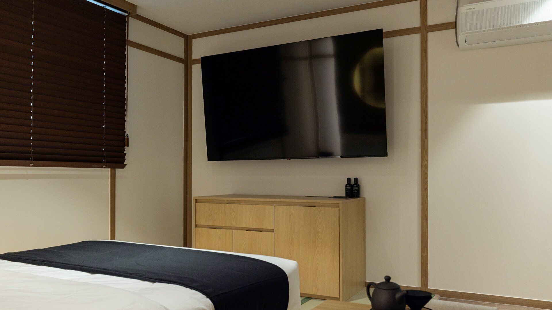 A hotel room with a bed, a TV mounted on the wall, a wooden cabinet, and a small table with a teapot.