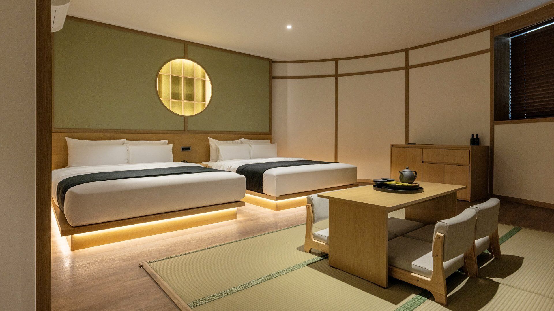 A hotel room with two beds, a small table, chairs, a cabinet, and a wall-mounted light fixture.