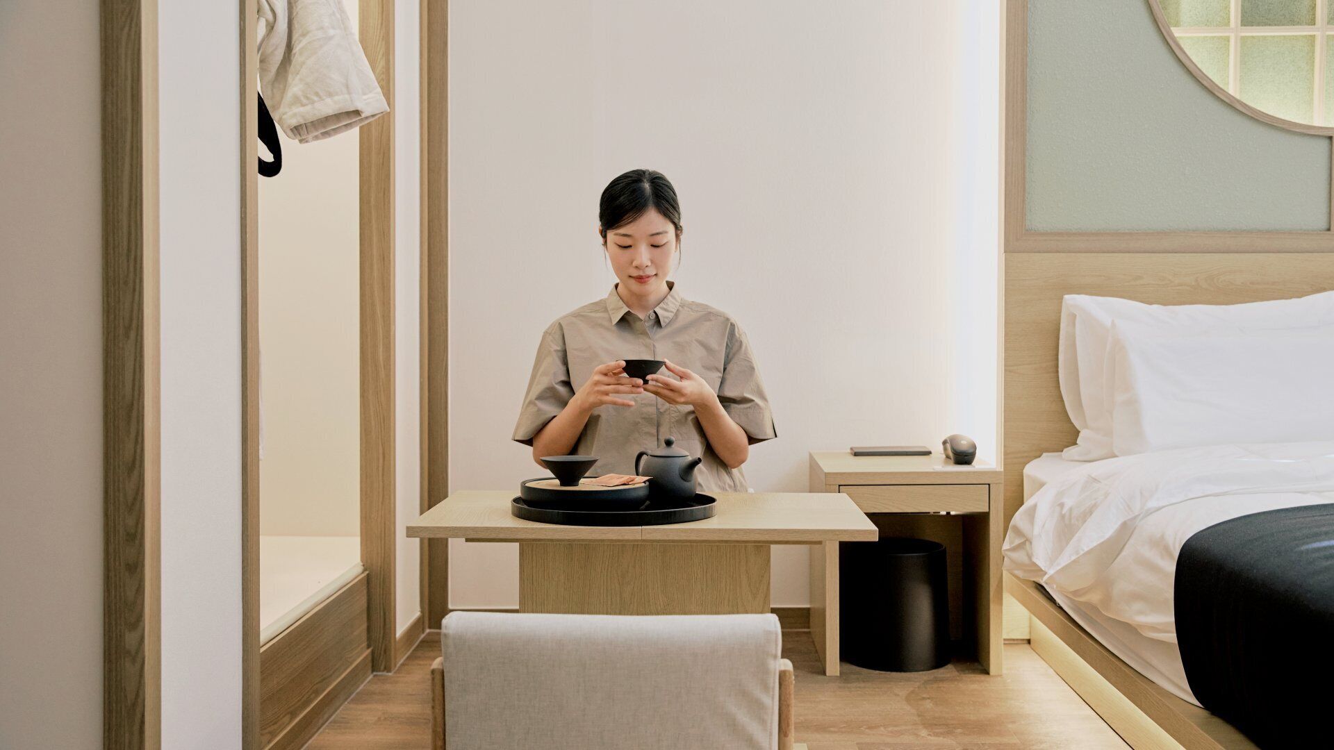 A hotel room with a bed, a bedside table with a lamp, a small table with a teapot and cups, and a trash can.