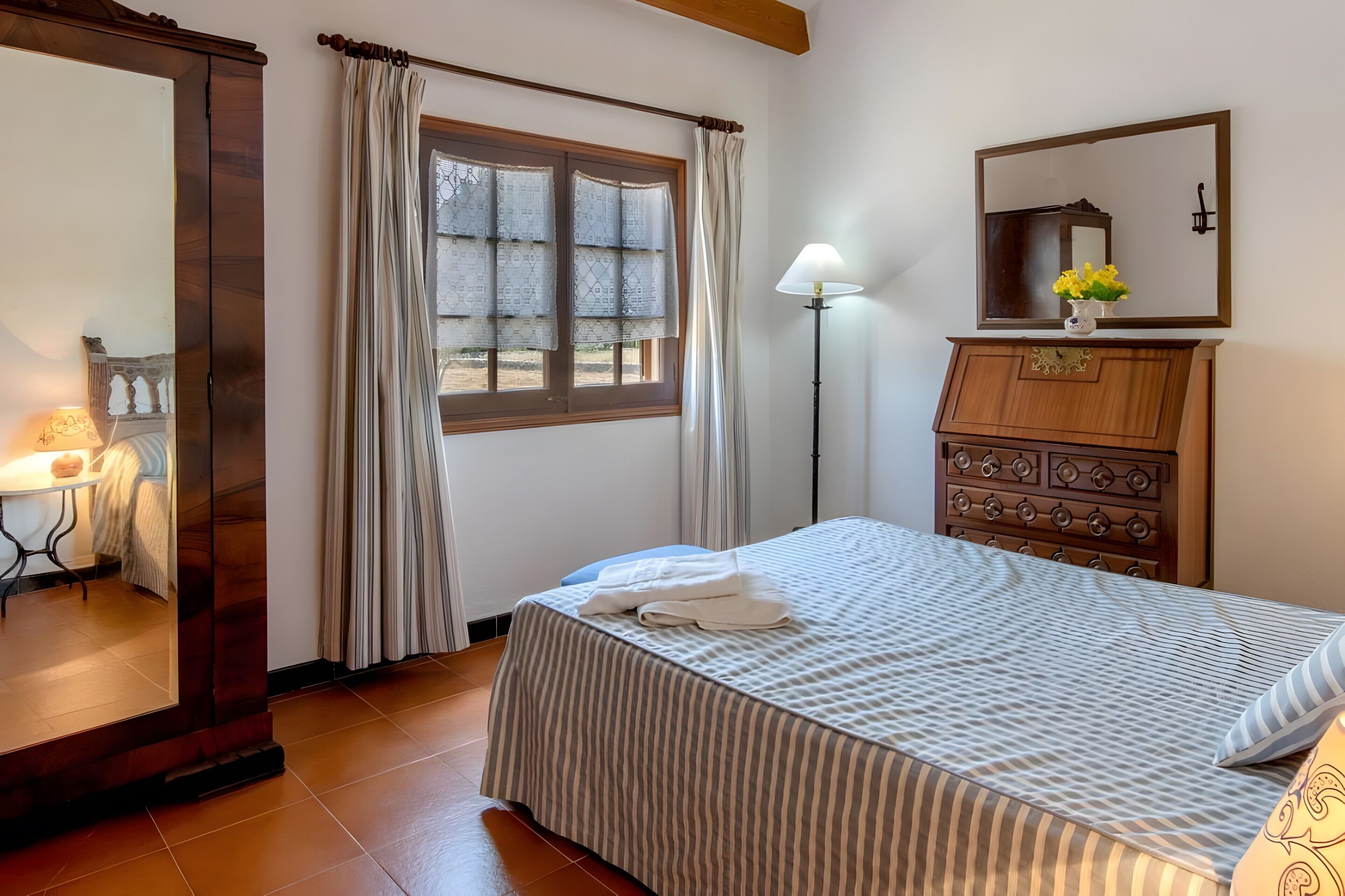 Une chambre à coucher avec un lit, une commode, un miroir, une fenêtre avec des rideaux et une lampe.