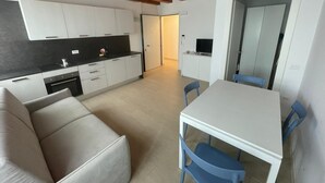 Eine moderne Küche mit weißen Schränken und einer schwarzen Arbeitsplatte, ein Essbereich mit weißem Tisch und blauen Stühlen sowie eine Couch.