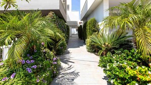 A pathway flanked by lush greenery and flowering plants, leading to a modern building with a flat roof and large windows. - Lemon Tree 1-BR Apt & Pool in Livadia (Larnaca)