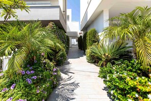 Un sendero bordeado por exuberante follaje y plantas florecientes, que conduce a un edificio moderno con tejado plano y grandes ventanales.