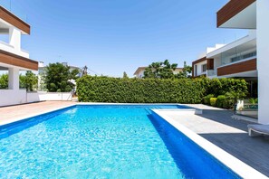 A swimming pool with a clear blue water surface, surrounded by modern buildings and a well-maintained hedge. - Lemon Tree 1-BR Apt & Pool in Livadia (Larnaca)