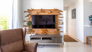 A modern living room with a flat-screen TV mounted on a wooden wall unit, a brown sofa, and a small side table with a plant. - Lemon Tree 1-BR Apt & Pool in Livadia (Larnaca)