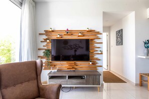 A modern living room with a flat-screen TV mounted on a wooden wall unit, a brown sofa, and a small side table with a plant. - Lemon Tree 1-BR Apt & Pool in Livadia (Larnaca)
