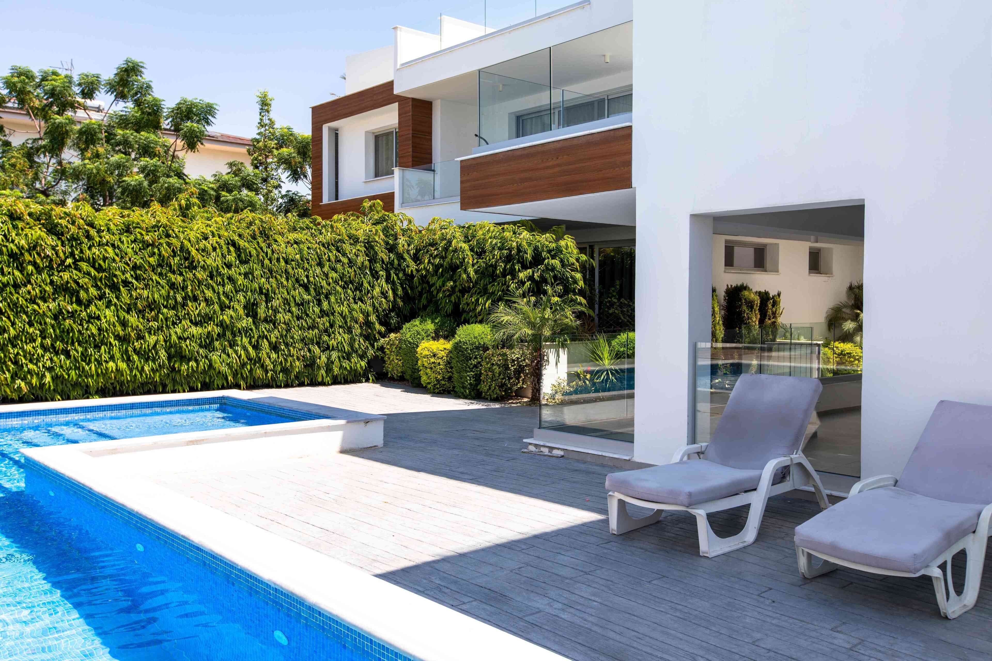 Un área de piscina moderna al aire libre con una terraza de madera, dos sillones de descanso y una barandilla de cristal.