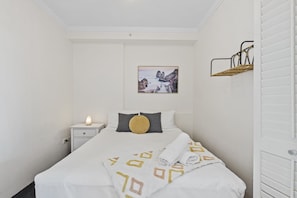 Cama bem arrumada com edredom branco e manta de padrão geométrico, luminária de cabeceira, mesa de cabeceira pequena, prateleira na parede e quadro com cena costeira.