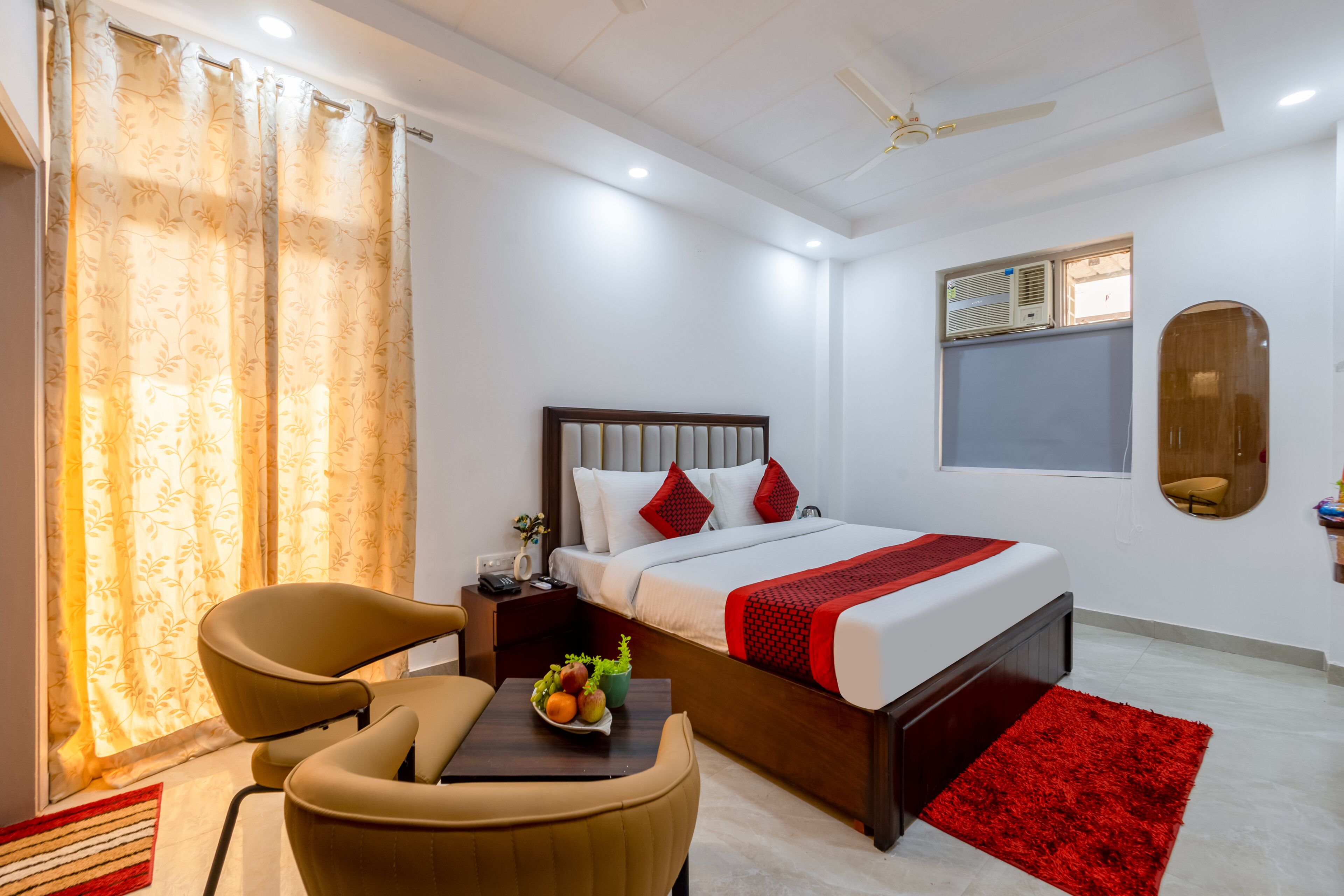 A hotel room with a bed, a desk, a chair, a red rug, and a window with curtains.