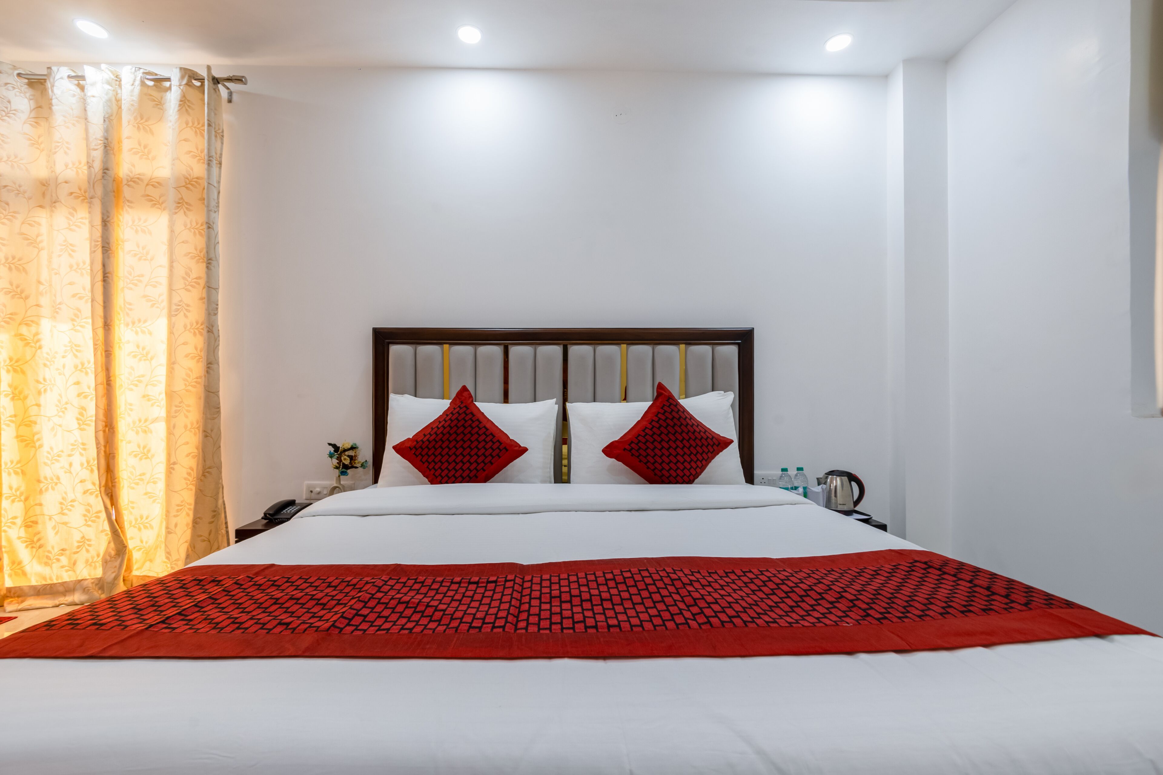A hotel room with a large bed, white bedding, two red pillows, a red blanket, a wooden headboard, a nightstand with a phone and a kettle, and a window with curtains.