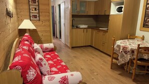A cozy room with a wooden sofa, a dining table with chairs, a kitchen area with wooden cabinets, and a wall-mounted lamp. - Les 2 alpes, at the foot of the slopes - apartments (Les Deux Alpes)