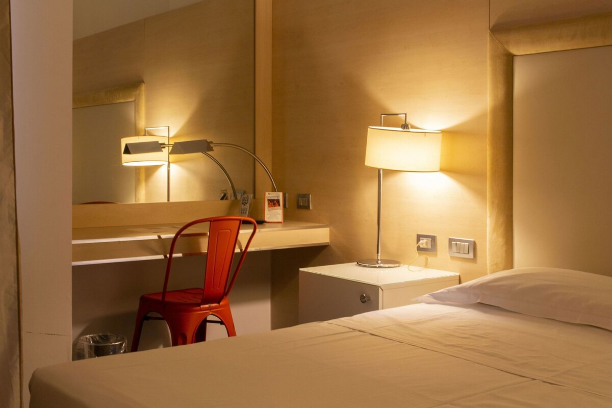 A hotel room with a bed, a chair, a desk, a lamp, a mirror, and a nightstand.