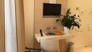 Una habitación con un escritorio, un televisor montado en la pared, una planta en maceta y un cubo de basura.