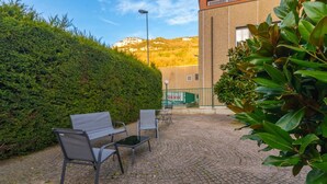 Un'area esterna pavimentata con una panchina e sedie, un siepe e un edificio sullo sfondo.