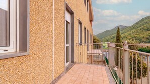 Un edificio con fachada de textura de guijarros, un balcón con barandilla de metal y montañas al fondo.