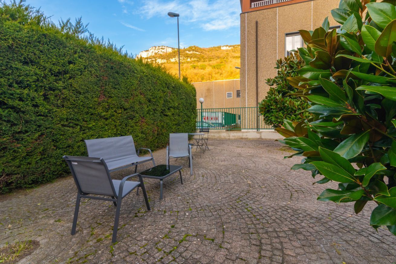 A paved outdoor area with a bench and chairs, a hedge, and a building in the background.