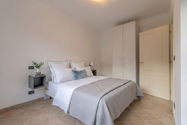 A bedroom with a bed, bedside tables, a nightstand, a door, and a wall-mounted shelf.