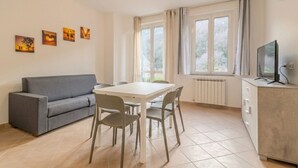 A room with a grey sofa, a white dining table with four chairs, a TV on a cabinet, and a window with curtains. - Piergi Bike Home 3 Finale Ligure (Finale Ligure)
