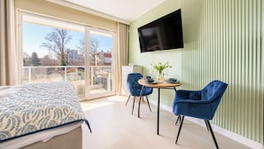 Une chambre moderne équipée d’un lit, d’une table à manger avec des chaises, d’une télévision et d’un balcon offrant une vue sur les bâtiments et les arbres.
