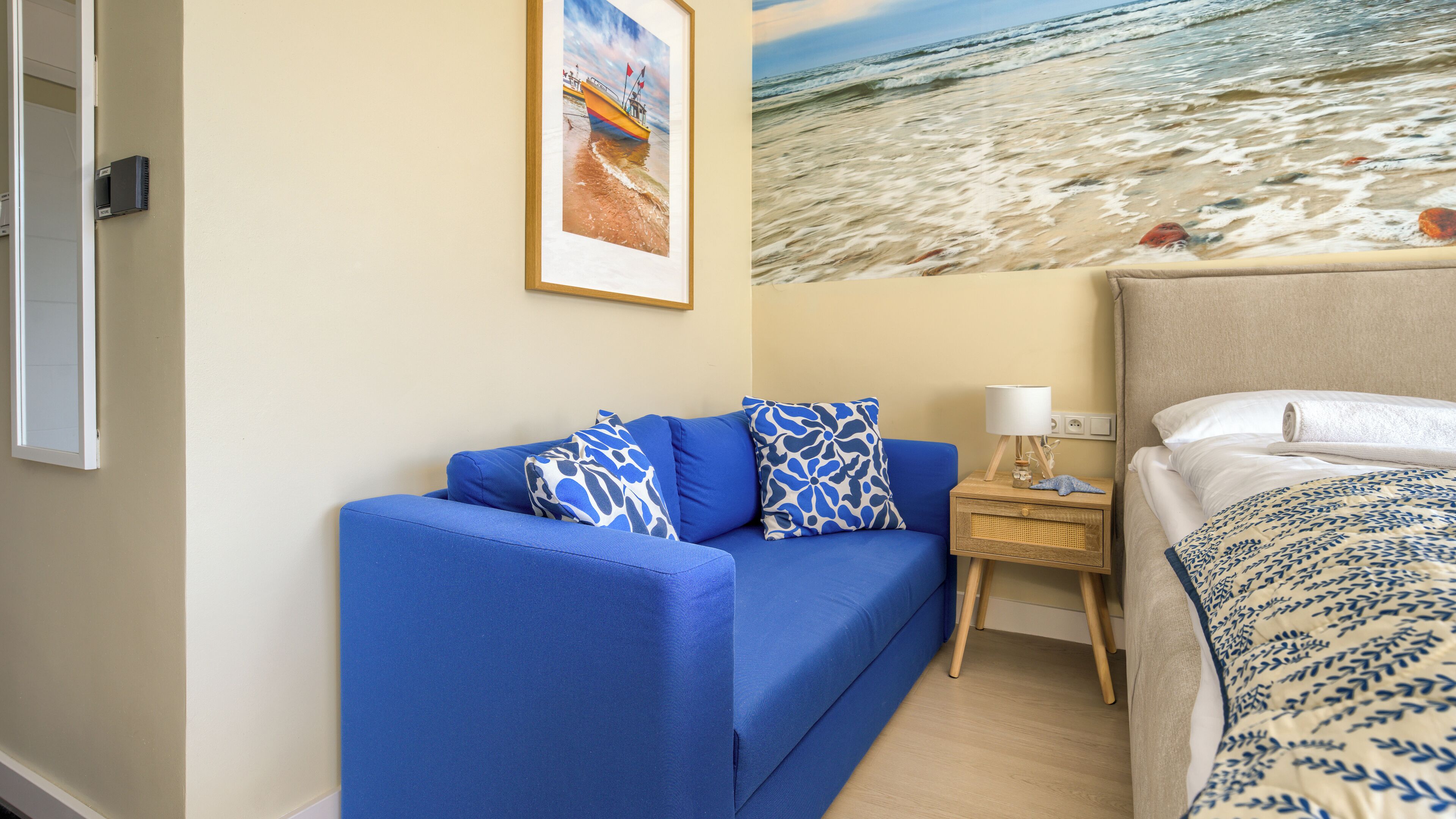 Ein Zimmer mit einem blauen Sofa, einem Bett mit gemustertem Überwurf, einem Nachttisch mit Lampe und einem gerahmten Bild von einer Strandlandschaft.