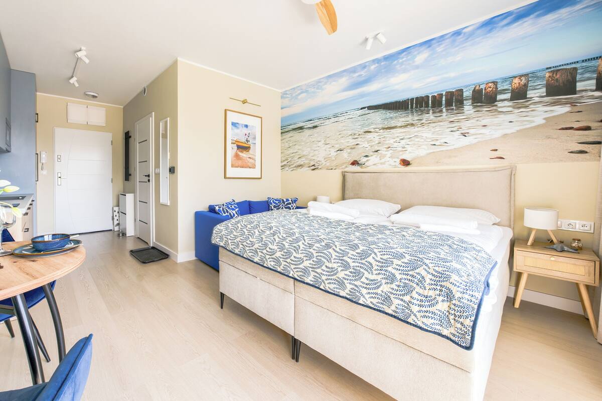 Une chambre à coucher avec un lit, un bureau agrémenté d’un vase de fleurs et un tableau représentant une scène de plage.