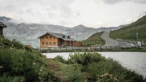 Un edificio de madera con un camino pavimentado que lo lleva a él, rodeado de montañas y una masa de agua.