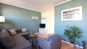 Ein Wohnzimmer mit grauem Sofa, einem kleinen Tisch mit Vase, einer Topfpflanze, einem Fernseher auf einem Schrank und einem gerahmten Bild an der Wand.