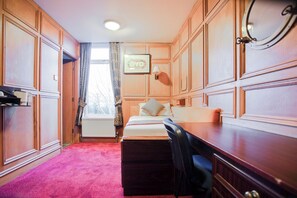Habitación de hotel con paneles de madera, una cama con almohadas, un escritorio, una silla, una ventana con cortinas y un cuadro en la pared.