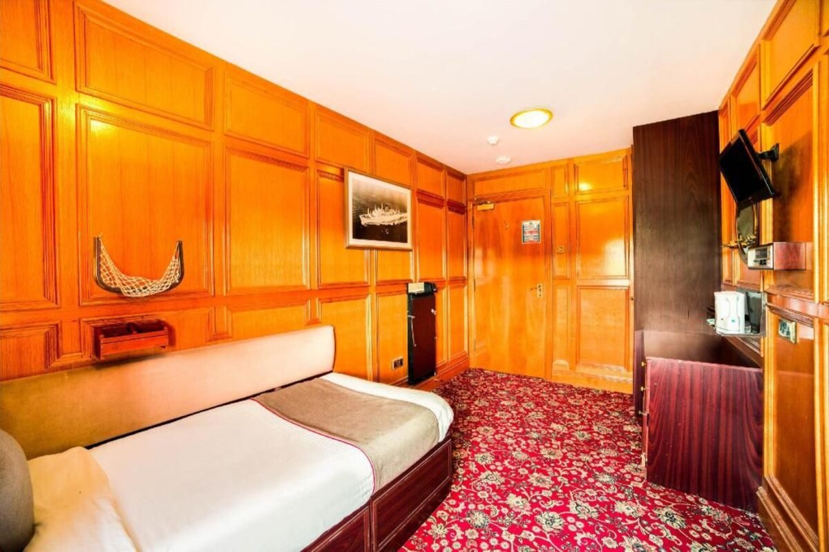 Een hotelkamer met een groot bed, houten lambrisering, een rood kamerkleed met dessin, een klein bureau en een televisie.