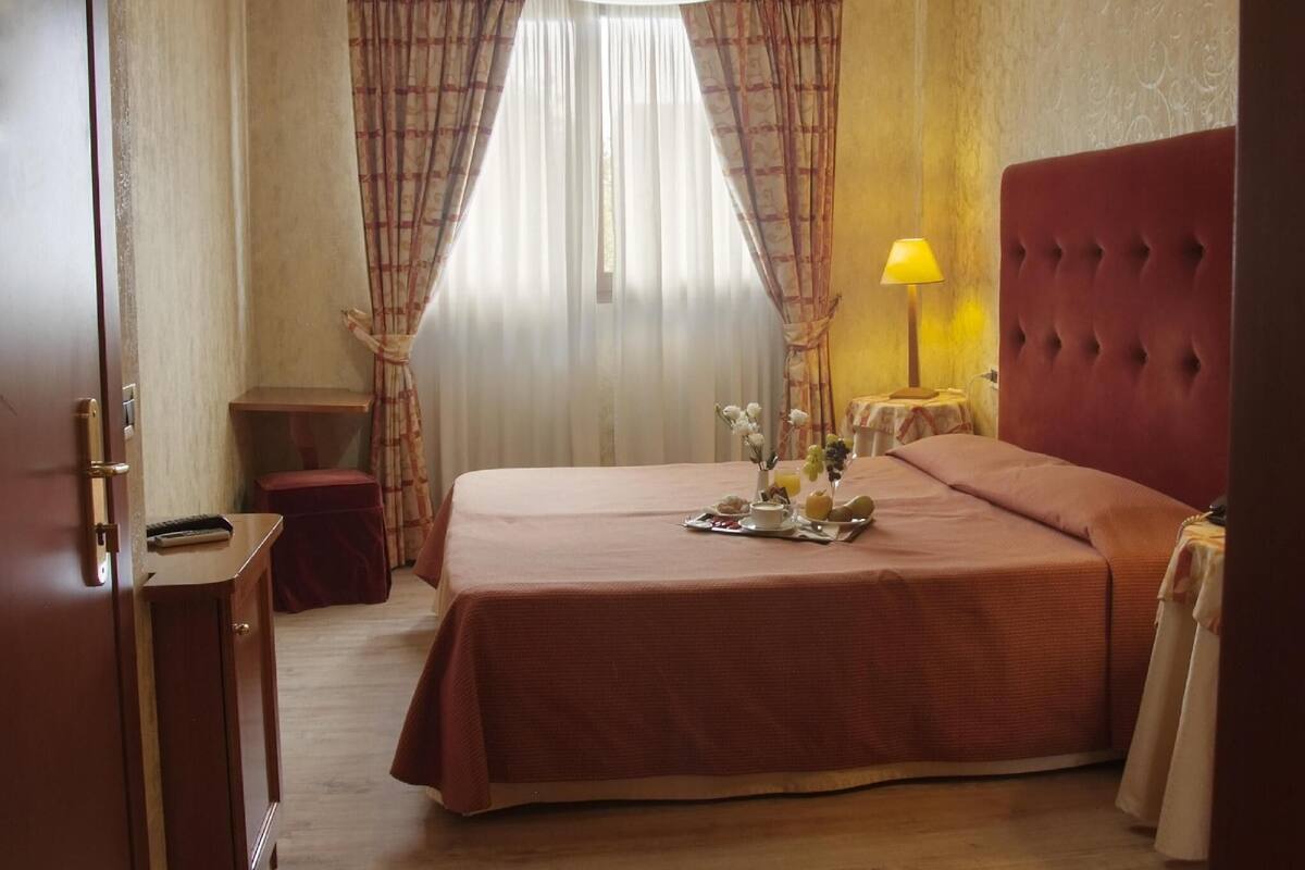 A hotel room with a bed, a bedside table, a lamp, curtains, and a window.